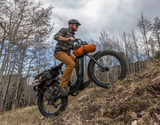 A cyclist wearing a helmet rides a Velotric Nomad 2X fat tire ebike uphill on a dirt trail with trees, demonstrating the "Power Unleashed" theme of the ebike